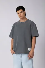Remeron de corte oversize color gris oscuro con efecto lavado. Presenta cuello redondo, mangas cortas con terminación desflecada y un parche rectangular con logo en la parte inferior frontal.