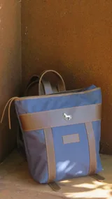 Mochila Sierra Mora confeccionada en lona azul con detalles en cuero marrón. Presenta un diseño estructurado con correas ajustables, cierre superior y un aplique metálico en forma de caballo en la parte frontal.