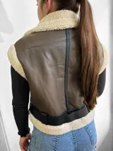 Chaleco marrón de cuero sintético con interior de piel sintética color beige. Tiene cierre frontal con cremallera, bolsillos laterales con cremallera y cinturón con hebilla.