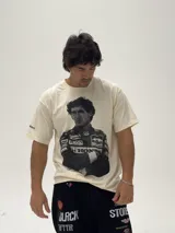 Remera de algodón color beige con estampa fotográfica frontal de Ayrton Senna con su traje de piloto de Fórmula 1. Presenta un corte oversize y cuello redondo.