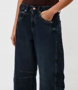 Pantalón de jean estilo baggy balloon, de tiro medio y corte amplio con diseño redondeado en las piernas. Presenta recortes laterales, cierre frontal con botón y cremallera, y bolsillos funcionales.