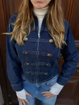 Chaqueta de denim azul oscuro con estilo militar, cuello alto, cierre frontal con cremallera y detalles de trabas decorativas con botones dorados.