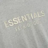 Camiseta gris con logo "Essentials Fear of God" en el pecho y etiqueta de goma en la parte posterior. Corte oversize.