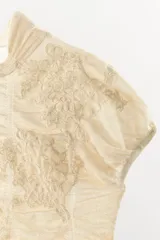 Top de punto con mezcla de viscosa, color beige, con cuello subido y manga corta. Presenta un diseño con frunces verticales y detalles bordados a tono, con terminación de ruedo ondulado.