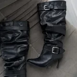 Botas altas negras de ecuero con diseño slouchy, tacón fino alto, puntera fina y tiras con hebillas metálicas ajustables en la caña y el tobillo.