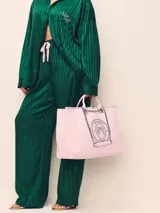 Bolso tipo shopper de Victoria's Secret, confeccionado en lona con diseño a rayas rosadas y blancas. Cuenta con asas de mano y correas de hombro con cadena metálica. Incluye un parche circular con el logo de la marca y alas bordadas en el centro.