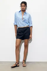 Bermuda de tiro medio color azul marino, con pinzas delanteras, cierre frontal y cinturón fino color beige.