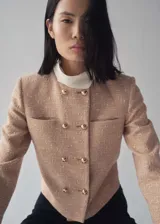 Chaqueta de tweed en color beige con diseño recto, cuello redondo y cierre frontal de doble botonadura con botones dorados. Presenta dos bolsillos de parche en el pecho y forro interior.