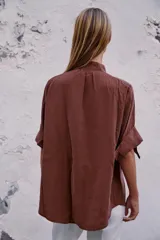 Camisa bordó de cuello alto ajustable con doble hebilla y cierre frontal con botones ocultos por solapa. Tiene mangas cortas con trabillas y diseño holgado.