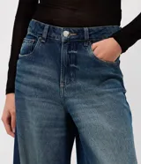 Pantalón de jean con corte super wide leg y tiro medio. Presenta un lavado uniforme con efecto levemente marcado, cintura con pasantes, cierre frontal con botón y cremallera, y bolsillos funcionales. Incluye remaches metálicos decorativos en forma de corazón.