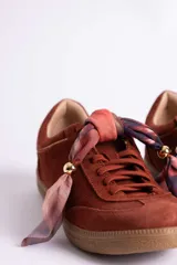 Zapatillas de gamuza color marrón con suela de goma color caramelo. Presentan cordones al tono y un detalle decorativo de pañuelo estampado atado en los laterales con esferas metálicas doradas.
