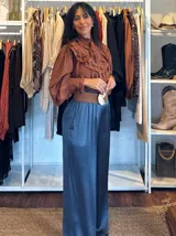 Blusa de color marrón con diseño romántico, cuello alto con lazo ajustable y volados tipo shabot en el frente. Presenta mangas largas abullonadas con puños ajustados y una textura suave y liviana.