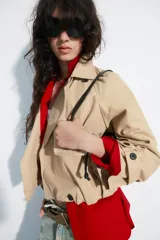 Trench corto de cuello solapa y escote en pico, color beige, con mangas largas terminadas en trabillas con botones. Cuenta con bolsillos laterales con solapa, cierre frontal cruzado con botón y bajo abullonado con elástico interior.