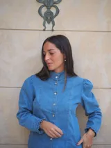 Camisa de jean de manga larga con cuello alto, cierre frontal de botones y hombros abullonados con fruncidos sutiles. Presenta terminaciones con volados delicados en los puños.