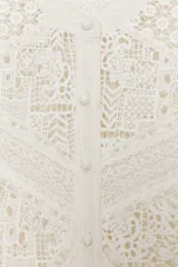 Blusa blanca de algodón con cuello redondo, manga larga con volante en el puño y detalle de bordados y aplicaciones de encaje en paneles geométricos. Cierre frontal con botones.