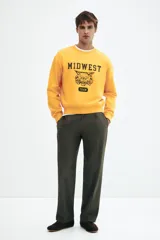 Buzo de tejido pesado de algodón color amarillo mostaza, con cuello redondo ribeteado, hombros caídos y puños y bajo en punto acanalado. Estampado frontal con el texto "MIDWEST" en mayúsculas, un dibujo de un felino rugiendo y la palabra "TEAM" en un óvalo.