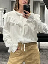 Camisa blanca de textura rugosa con volados en el pecho y hombros, cuello redondo con lazos de ajuste, cierre frontal con botones y mangas largas con puños con volados.