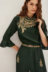 Camisa de viscosa color verde oscuro, con cuello alto y mangas tres cuartos terminadas en volados. Presenta un elaborado bordado artesanal en el pecho con pedrería dorada y detalles decorativos en la cintura.