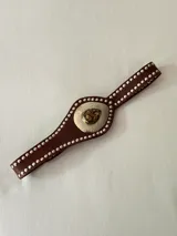 Cinto faja de cuero marrón con tachas metálicas plateadas en los bordes. Presenta una pieza central metálica con textura martillada y un dije en forma de gota con piedra natural.