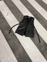 Chaleco de cuero negro, abierto a los costados, con tiras que se regulan a necesidad y gusto de cada una. Presenta bolsillos frontales y escote en V.