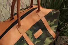 Bolso de lona con estampado de camuflaje y detalles en cuero color suela. Tiene doble asa corta de cuero y cierre superior con cremallera.