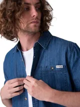 Camisa de manga corta en denim ligero de color azul, con cierre frontal de botones, cuello camisero y un bolsillo en el pecho con solapa y botón.