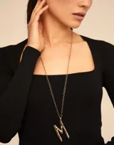 Dije con forma de letra M, bañado en oro, con diseño inspirado en un clavo. Pieza artesanal de estilo minimalista.