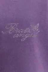 Camiseta de manga larga con corte oversize, confeccionada en algodón color violeta con efecto lavado. Presenta cuello redondo, bajo asimétrico y detalles de texto con aplicación de brillos en el pecho y las mangas.