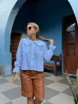 Camisa blanca de corte holgado con cuello bebé con volados, cierre frontal de botones y mangas largas con puños terminados en volados.