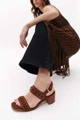 Sandalias de cuero color marrón con dos tiras horizontales, una sobre los dedos y otra sobre el empeine, ambas decoradas con tachas. Tienen taco cuadrado de madera y cierre con hebilla.