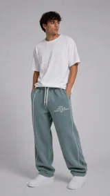 Pantalón de jogging color verde azulado, de corte holgado y diseño deportivo. Presenta líneas verticales blancas a los costados, cintura elástica con cordón ajustable y estampado tipográfico en la parte frontal.
