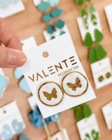 Caravanas circulares con borde dorado, fondo blanco texturizado y aplique central de mariposa dorada.