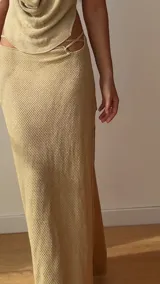 Falda larga tejida color marrón con brillo metálico, corte al cuerpo, abertura alta en la pierna y recortes laterales con cordones ajustables.