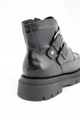 Bota corta de cuero negro con suela track dentada y dos correas decorativas con hebillas metálicas en el lateral. Presenta un diseño de puntera cuadrada y cierre lateral.