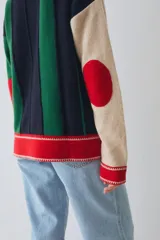 Buzo estilo polo de lambswool con diseño color block a rayas verticales en tonos beige, marrón y rojo. Presenta cuello camisero, cartera frontal con botones, mangas largas con coderas y tajos laterales en el ruedo.