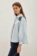 Camisa blanca de algodón con cuello camisero y mangas largas abullonadas con puños ajustados. Presenta detalles bordados en los hombros con motivos vegetales y aplicaciones brillantes.