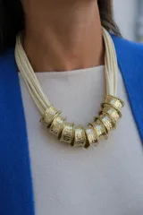 Collar de diseño artesanal compuesto por múltiples tiras de cuero en color crudo y piezas cilíndricas de metal dorado con textura martillada.