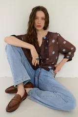 Blusa marrón semitransparente con estampado de lunares celestes, mangas largas abuchonadas, cuello en V con lazos y detalles de volados frontales.
