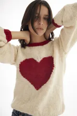 Buzo de tejido de punto artesanal, color crema con un gran corazón negro estampado en el centro y detalles de franjas negras en las mangas. Presenta cuello redondo y puños acanalados.