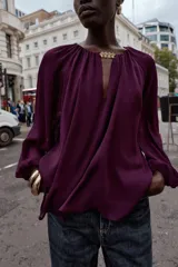 Blusa de tejido semitransparente color violeta, con cuello redondo y detalle de fornitura dorada en el escote. Presenta un diseño de escote en pico, mangas largas abullonadas con puños elásticos y corte holgado.