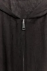 Cazadora semitransparente color marrón oscuro, con cuello redondo, capucha y manga larga abullonada. El bajo y los puños tienen acabado elástico. Cierre frontal con cremallera metálica.