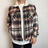 Chaqueta de estilo leñador con estampado de cuadros en tonos marrones, blancos y negros. Presenta cuello de corderito, cierre frontal con botones, bolsillos en el pecho con solapa y bolsillos laterales.