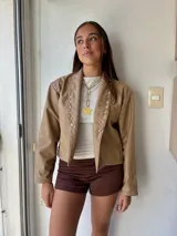Campera de ecocuero color beige, con diseño de solapas decoradas con tachas metálicas plateadas y cierre frontal abierto.