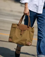Cartera tipo tote bag color marrón con detalles en cuero sintético tostado, solapa frontal con tira decorativa, doble asa de mano y refuerzos en las esquinas inferiores.
