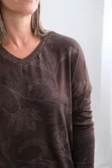 Sweter de punto color chocolate con cuello en V y diseño estampado con motivos botánicos en tonos más claros. Presenta mangas largas y un corte holgado con terminación acanalada en el ruedo.