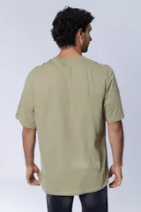 Remera oversize color verde oliva con cuello redondo, mangas cortas y un parche cuadrado negro con un logo en el pecho. Presenta un estampado de texto en el lado izquierdo del pecho.