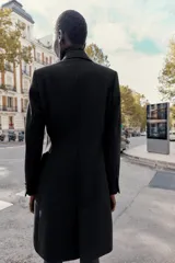 Abrigo de paño color beige con cuello solapa, manga larga con hombreras y cierre frontal con botones. Presenta bolsillos delanteros de doble vivo y abertura en la espalda.