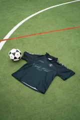 Camiseta de fútbol estilo boxy de manga corta y cuello polo, en color negro con sutiles rayas verticales. Presenta inscripciones estampadas y escudo bordado en el pecho.