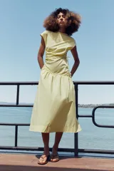 Vestido midi confeccionado en hilatura de algodón de color amarillo, con cuello redondo, manga corta y detalles de frunces laterales ajustables con cordones. Presenta bajo asimétrico con acabado en línea evasé.