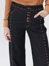 Jeans de corte ancho (wide leg) y tiro medio, confeccionados en denim de algodón negro con costuras contrastantes en color ocre. Presenta múltiples botones metálicos frontales y tachas metálicas aplicadas a lo largo de los laterales y la pretina.